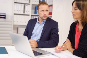 Worried businessman listening to a middle-aged female colleague as they sit together at a table in the office sharing a laptop computer